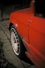 A close-up view of a red car with a focus on its rear fender and a white wheel featuring red text that reads 'OZ Racing'. The car's bodywork shows signs of damage near the wheel arch. The background is dimly lit and out of focus, suggesting nighttime.