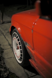 A close-up view of a red car with a focus on its rear fender and a white wheel featuring red text that reads 'OZ Racing'. The car's bodywork shows signs of damage near the wheel arch. The background is dimly lit and out of focus, suggesting nighttime.