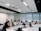 A group of people are seated in a modern classroom or lecture hall setting. Most are using laptops, indicating a possible workshop or educational session. The room has a tiered seating arrangement with a white interior and large windows at the back. There are projectors mounted on the ceiling.