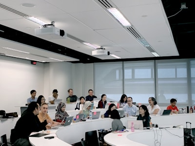 A group of people are seated in a modern classroom or lecture hall setting. Most are using laptops, indicating a possible workshop or educational session. The room has a tiered seating arrangement with a white interior and large windows at the back. There are projectors mounted on the ceiling.