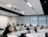 A group of people are seated in a modern classroom or lecture hall setting. Most are using laptops, indicating a possible workshop or educational session. The room has a tiered seating arrangement with a white interior and large windows at the back. There are projectors mounted on the ceiling.