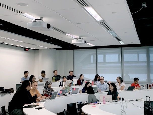 A group of people are seated in a modern classroom or lecture hall setting. Most are using laptops, indicating a possible workshop or educational session. The room has a tiered seating arrangement with a white interior and large windows at the back. There are projectors mounted on the ceiling.