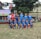 A group of young boys in soccer uniforms are posing on a grassy field. Most of them are wearing blue jerseys, while one wears a red jersey. They are in front of a backdrop of trees and buildings. A banner in the background suggests this is a sports or soccer event.