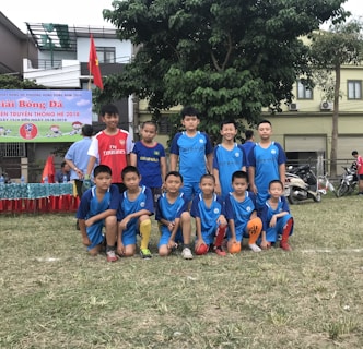 A group of young boys in soccer uniforms are posing on a grassy field. Most of them are wearing blue jerseys, while one wears a red jersey. They are in front of a backdrop of trees and buildings. A banner in the background suggests this is a sports or soccer event.