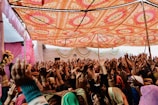 A lively celebration is taking place under a vibrant, intricately patterned canopy. Numerous people are gathered closely together, raising their hands in joyful participation. The setting is decorated with pink balloons and floral arrangements, creating a festive atmosphere.