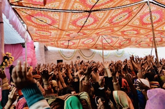 A lively celebration is taking place under a vibrant, intricately patterned canopy. Numerous people are gathered closely together, raising their hands in joyful participation. The setting is decorated with pink balloons and floral arrangements, creating a festive atmosphere.