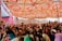 A lively celebration is taking place under a vibrant, intricately patterned canopy. Numerous people are gathered closely together, raising their hands in joyful participation. The setting is decorated with pink balloons and floral arrangements, creating a festive atmosphere.