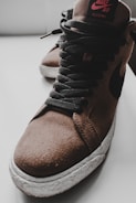 A close-up of a brown suede high-top sneaker with black laces and a textured white sole. The shoe features a prominent swoosh design and branding on the tongue.