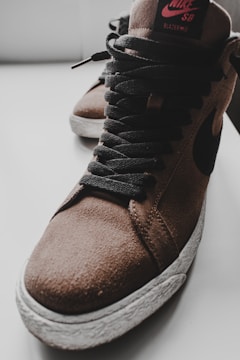 A close-up of a brown suede high-top sneaker with black laces and a textured white sole. The shoe features a prominent swoosh design and branding on the tongue.