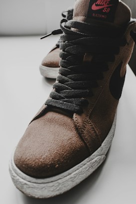 A close-up of a brown suede high-top sneaker with black laces and a textured white sole. The shoe features a prominent swoosh design and branding on the tongue.