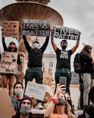 A group of people participating in a protest, holding signs with messages like 'Black Lives Matter' and 'Time to change the World.' Some participants are wearing face masks, and a large stone fountain is visible in the background.