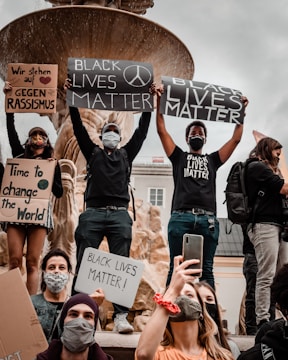 A group of people participating in a protest, holding signs with messages like 'Black Lives Matter' and 'Time to change the World.' Some participants are wearing face masks, and a large stone fountain is visible in the background.