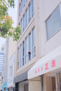 A multi-story building with large windows and light-colored facade. A person is partially visible, leaning out of one of the windows, appearing to hold something. A tree with green leaves is in the foreground, and there are various signs in Japanese and awnings with Japanese text.