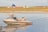 A jet ski is floating on a calm body of water with grassy banks in the background. In the distance, there is a large inflatable water slide and a row of small, wooden cabins amidst colorful buildings.