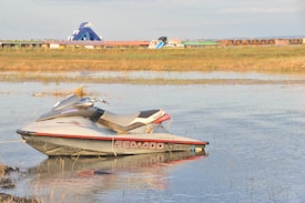 A jet ski is floating on a calm body of water with grassy banks in the background. In the distance, there is a large inflatable water slide and a row of small, wooden cabins amidst colorful buildings.