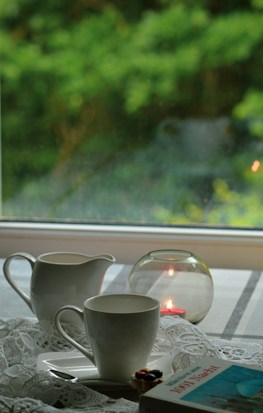 A serene indoor scene with a white ceramic cup and creamer, a small burning candle inside a glass holder, a partially visible book, and lace fabric on the table. The background shows a window revealing lush green foliage outside, softly out of focus, enhancing the calm atmosphere.
