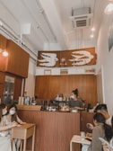 A cozy coffee shop interior with warm wooden decor where a barista prepares drinks behind the counter. Several patrons are seated, some engaged with their phones and others chatting. A decorative banner with stylized bird designs hangs above the counter.
