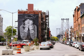 A vibrant urban street scene features a large mural of a contemplative person with striped clothing on the side of a brick building. A bridge stretches across in the background, while parked cars line the street. The area is bustling with brick buildings, some with fire escapes, and various street signs are visible.