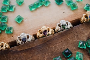 A collection of ornate rings featuring heart-shaped gemstones are arranged on a wooden surface. The rings display intricate designs with a mix of silver and gold metalwork, and each contains a differently colored gemstone set in the center. Scattered around the rings are several loose, square-cut green gemstones, possibly emeralds, adding a touch of vibrant color to the composition.