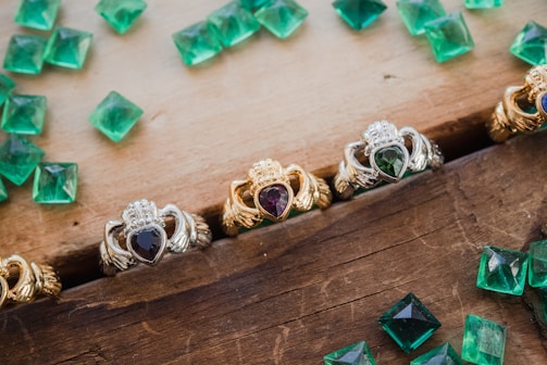 A collection of ornate rings featuring heart-shaped gemstones are arranged on a wooden surface. The rings display intricate designs with a mix of silver and gold metalwork, and each contains a differently colored gemstone set in the center. Scattered around the rings are several loose, square-cut green gemstones, possibly emeralds, adding a touch of vibrant color to the composition.