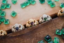 A collection of ornate rings featuring heart-shaped gemstones are arranged on a wooden surface. The rings display intricate designs with a mix of silver and gold metalwork, and each contains a differently colored gemstone set in the center. Scattered around the rings are several loose, square-cut green gemstones, possibly emeralds, adding a touch of vibrant color to the composition.
