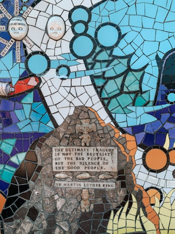 A colorful mosaic with various abstract and figurative elements, including two sculpted faces and alphabetical tiles with words like 'love', 'hope', and 'passion'. The focal point is a quote by Martin Luther King on a darker stone-like section surrounded by ornamental designs. The mosaic incorporates a diverse palette of blues, oranges, and browns, with intricate patterns resembling waves, celestial bodies, and organic shapes.