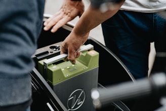 A person is seen inserting or removing a green battery from a device or vehicle, using both hands. The setting appears to involve a mechanical or electronic component.