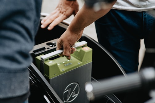 A person is seen inserting or removing a green battery from a device or vehicle, using both hands. The setting appears to involve a mechanical or electronic component.