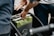A person is seen inserting or removing a green battery from a device or vehicle, using both hands. The setting appears to involve a mechanical or electronic component.