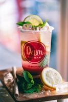 A refreshing beverage in a clear plastic cup, garnished with a lemon slice and mint leaves. The cup has a red logo label and is filled with ice cubes. It is placed on a wooden surface with a paper straw and more mint and lemon slices nearby.