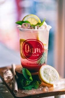 A refreshing beverage in a clear plastic cup, garnished with a lemon slice and mint leaves. The cup has a red logo label and is filled with ice cubes. It is placed on a wooden surface with a paper straw and more mint and lemon slices nearby.