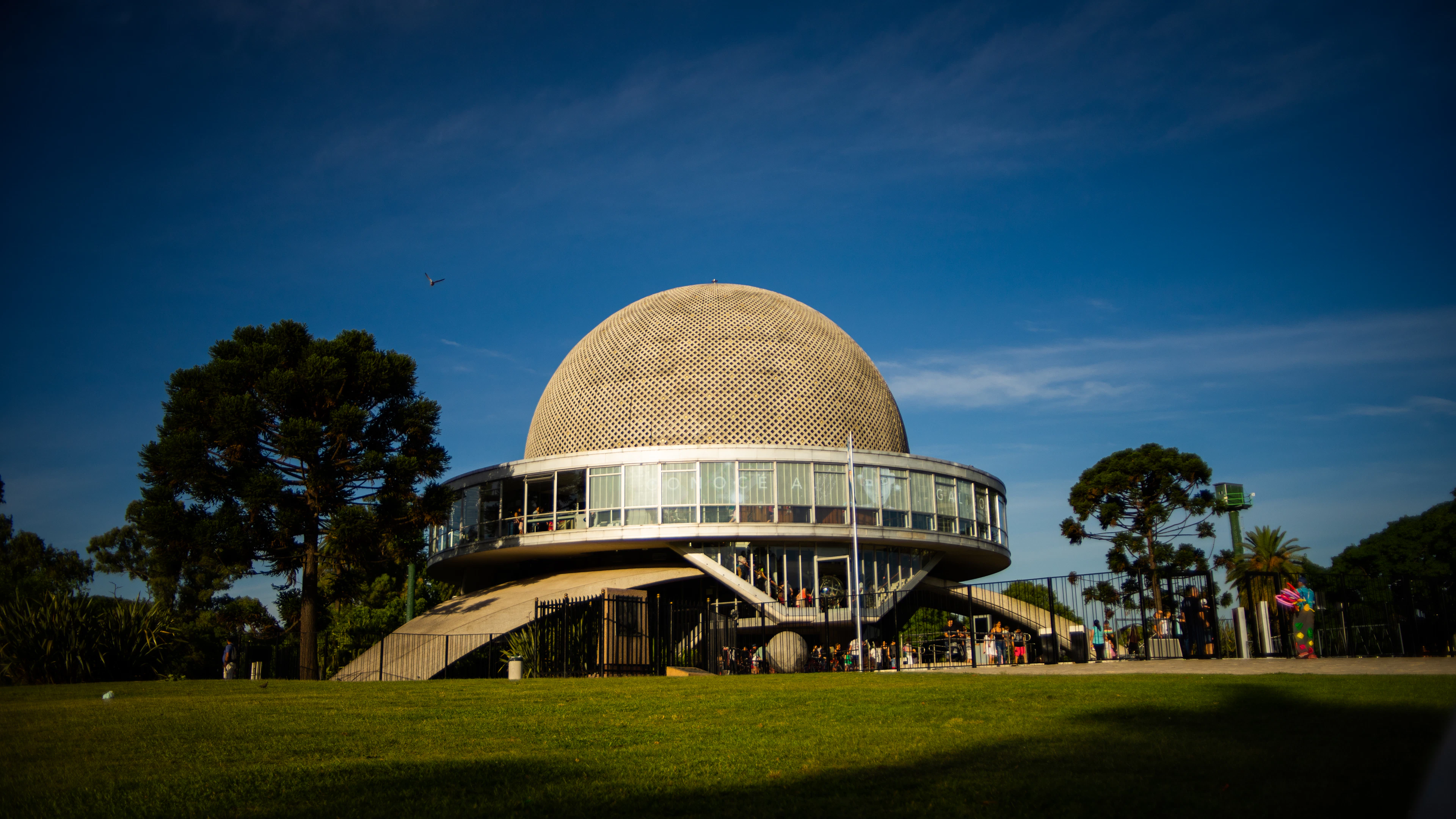 The flying-saucer dome of the Planetario Galileo Galilei in the Palermo parks, its perforated sphere resting on three concrete legs and lit warm against a deep blue evening sky above the lawn.