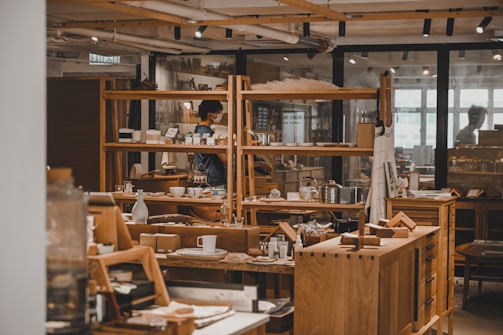 A cozy interior of a shop featuring wooden furniture and a variety of home goods, such as cups, books, and decorative items. Shelving units display an assortment of items, and a person wearing a mask can be seen browsing. Warm lighting enhances the inviting atmosphere.