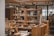 A cozy interior of a shop featuring wooden furniture and a variety of home goods, such as cups, books, and decorative items. Shelving units display an assortment of items, and a person wearing a mask can be seen browsing. Warm lighting enhances the inviting atmosphere.