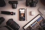 A variety of photography equipment and electronic devices are organized on a white surface. Items include two cameras, camera lenses, a phone displaying an image, a tablet with photographs, a digital pen, wireless earbuds, and a small tripod.