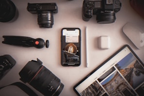 A variety of photography equipment and electronic devices are organized on a white surface. Items include two cameras, camera lenses, a phone displaying an image, a tablet with photographs, a digital pen, wireless earbuds, and a small tripod.