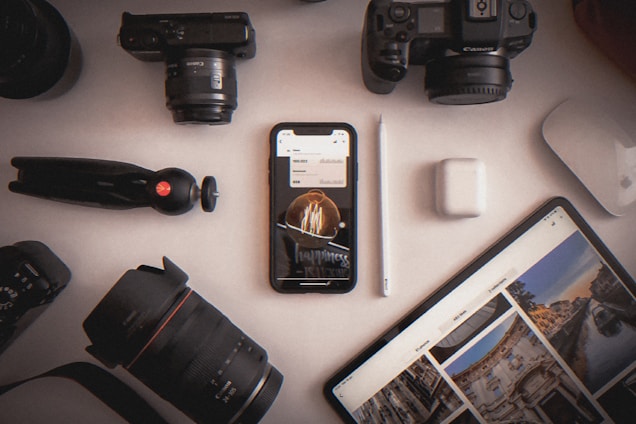 A variety of photography equipment and electronic devices are organized on a white surface. Items include two cameras, camera lenses, a phone displaying an image, a tablet with photographs, a digital pen, wireless earbuds, and a small tripod.