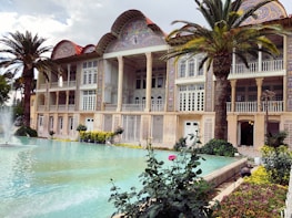 A beautifully ornate building with intricate tile work and archways is surrounded by lush palm trees and blooming flowers. A serene pool with fountains adds to the tranquil atmosphere.