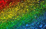 Vibrant splashes of paint create a gradient effect starting from red at the top left, blending through yellow and green, and transitioning to blue at the bottom right on a dark background. The droplets and splatters add a dynamic and chaotic feeling to the artwork.