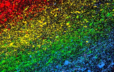 Vibrant splashes of paint create a gradient effect starting from red at the top left, blending through yellow and green, and transitioning to blue at the bottom right on a dark background. The droplets and splatters add a dynamic and chaotic feeling to the artwork.