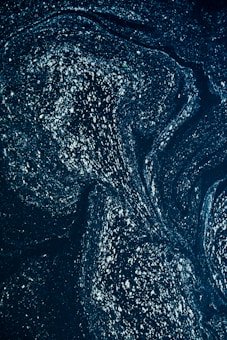A swirling pattern of shimmering specks on a dark background, resembling a night sky filled with stars or a galaxy viewed from afar. The image has a cosmic, abstract quality, with the bright specks creating a sense of depth and movement.