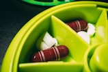 A close-up view of pills and capsules placed inside a circular green container with multiple compartments. The container is made of a plastic material and is designed for organizing medication. The pills include two large red and white capsules and several small white round tablets, all neatly arranged within the sections.