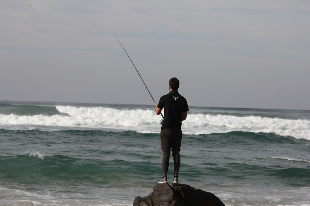 uomo in giacca nera e pantaloncini neri che tiene la canna da pesca in piedi sulla roccia marrone vicino al mare