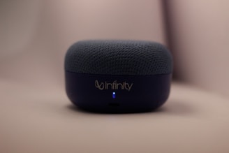 A small, dark blue audio speaker rests on a smooth surface with the brand name 'Infinity' on the front. The top is covered with a textured fabric material, and a small blue LED light is visible.