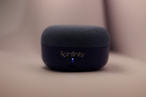 A small, dark blue audio speaker rests on a smooth surface with the brand name 'Infinity' on the front. The top is covered with a textured fabric material, and a small blue LED light is visible.