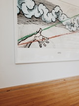 A framed artwork featuring a hand reaching out towards abstract, swirling cloud forms. The artwork is bordered by green and red lines. It is mounted on a white wall above a wooden floor.