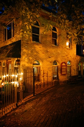 A brick building with arched windows is illuminated by warm lighting. Leafy branches hang over the top, adding a natural element. Lights are strung along a black metal fence, casting a cozy glow. The cobblestone pathway enhances the vintage and welcoming atmosphere.