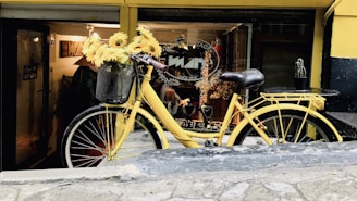 A vibrant yellow vintage bicycle is parked outside a shop with a basket full of sunflowers on its front. The storefront features a variety of objects, including jewelry displayed in the window. A small cactus in a pot is placed on the rear rack of the bicycle. The shop has a cozy atmosphere with warm lighting visible through the glass.