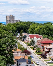A suburban neighborhood with rows of houses featuring diverse architectural styles, surrounded by lush green trees. In the background, a tall apartment building rises against a backdrop of distant hills and a sky filled with fluffy clouds.