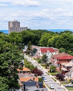 A suburban neighborhood with rows of houses featuring diverse architectural styles, surrounded by lush green trees. In the background, a tall apartment building rises against a backdrop of distant hills and a sky filled with fluffy clouds.